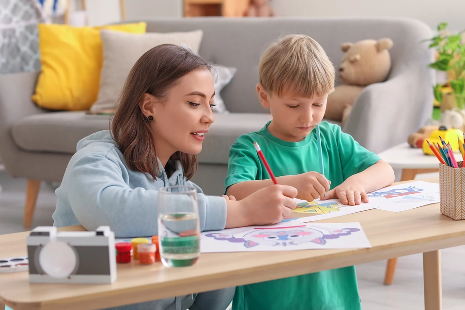 Une femme adulte accompagne un jeune garçon blond dans une activité de dessin et coloriage à domicile